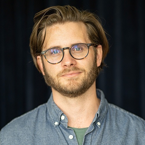 Portrait photo of Tobias Nyhuus Jensen, a man in his late thirties. He has a beard and brown, slightly unruly hair, brushed back and, according to his mother, in need of a haircut. He wears round glasses and a light blue shirt. His smile is friendly and slightly restrained, like a modern version of the Mona Lisa from the software industry—if she could grow stubble.