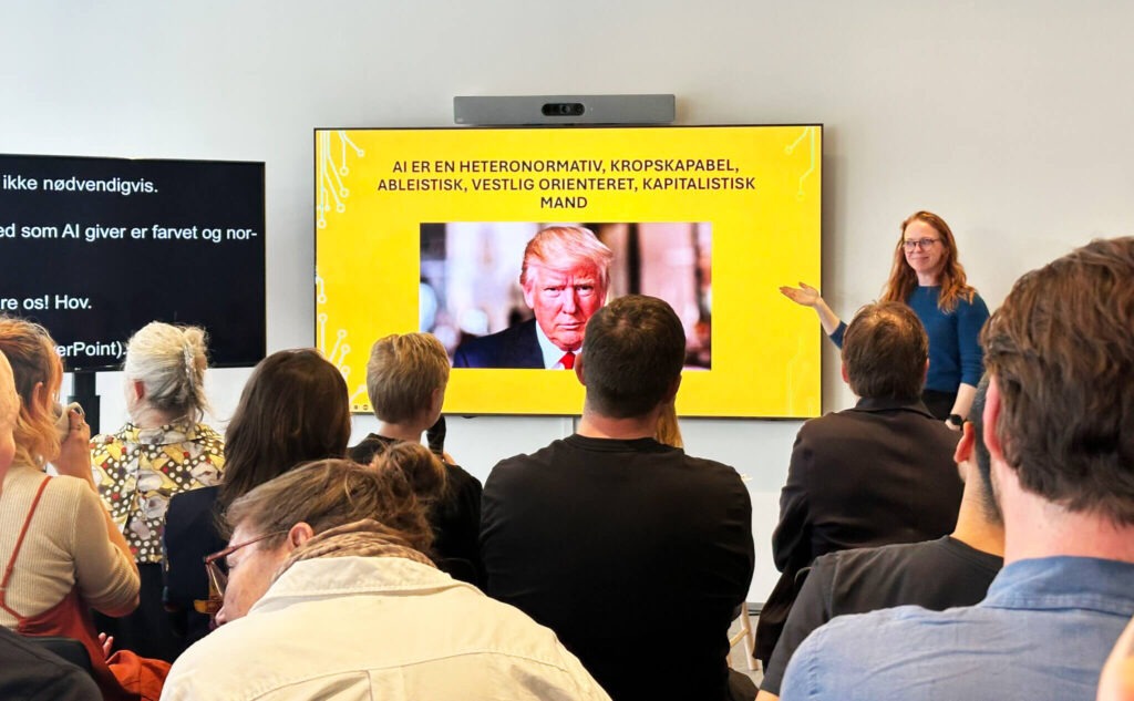 Presentation by Cathrine Mejdal, standing in front of a yellow slide with the text “AI is a heteronormative, body-capable, ableist, Western-oriented, capitalist man”. The audience follows along as live subtitles are displayed on the screen beside her.