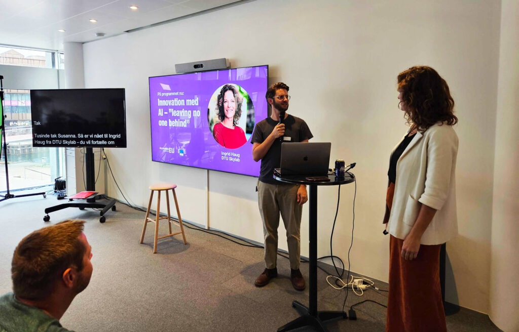 Ingrid Haug from DTU Skylab being introduced on stage in front of a slide titled “Innovation with AI – ‘leaving no one behind’”, while live subtitles are displayed on a screen beside her.