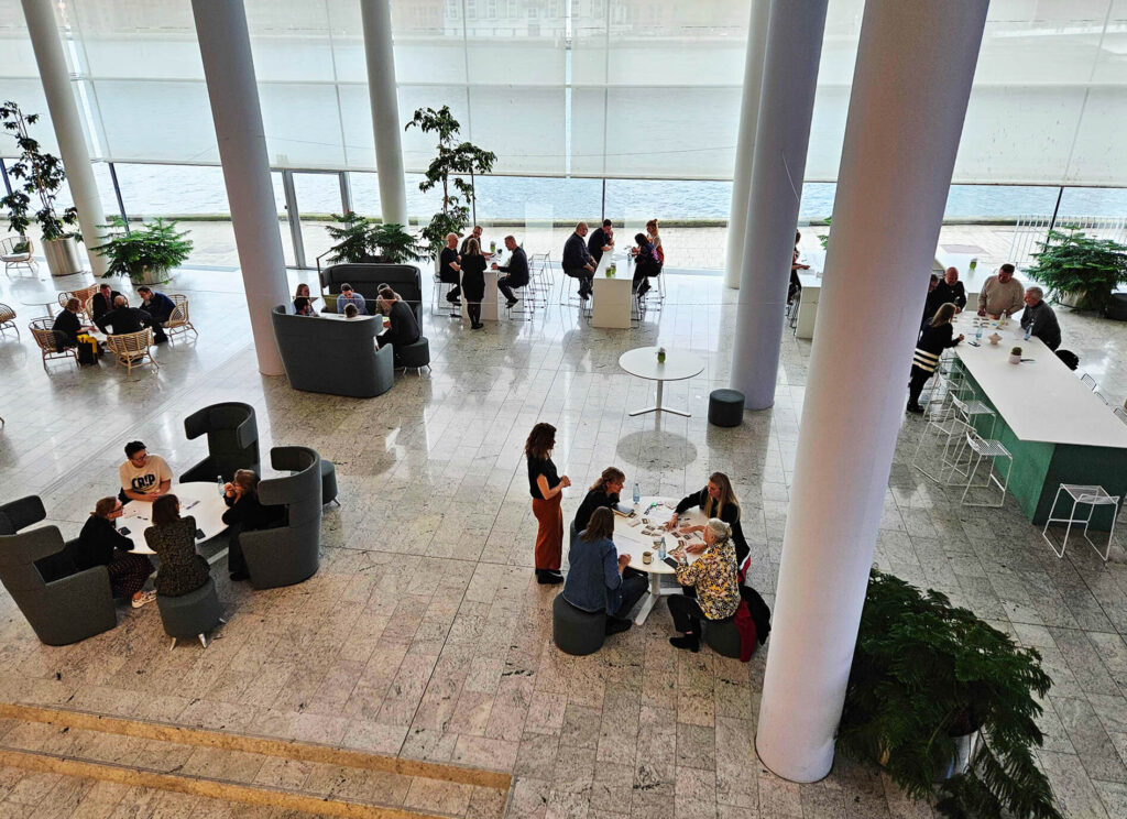 Aerial photo of the workshop led by Ingrid Haug from DTU Skylab, where participants work in small groups in a bright atrium with a view of the water.