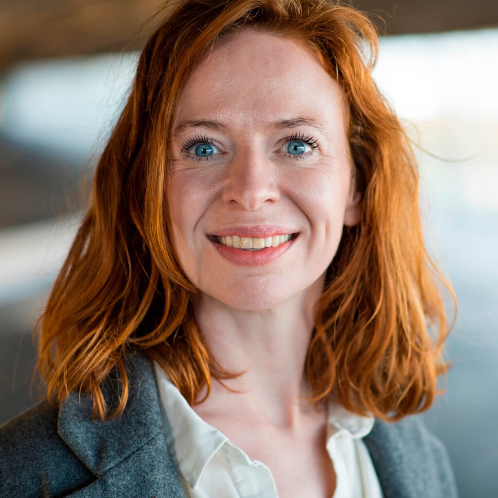Portrait of a woman with shoulder-length red hair and blue eyes. She is smiling and wearing a white blouse and a gray blazer, standing in a softly lit indoor or semi-outdoor environment with a blurred background.