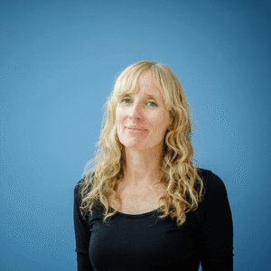 Portrait of Dorte Nørregaard, a woman with long, wavy blonde hair and blue eyes. She is wearing a black blouse and stands in front of a solid blue background.