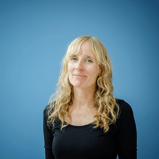 Portrait photo of a woman with long, light, curly hair wearing a black blouse, standing in front of a blue background.