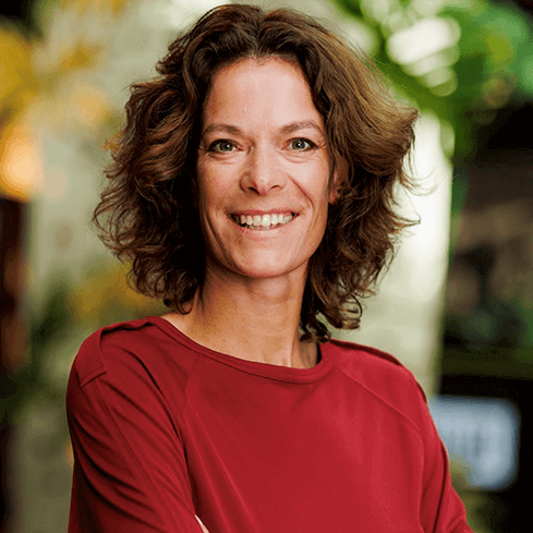 Portrait of a woman with medium-length, curly brown hair and brown eyes. She is smiling and wearing a red blouse, standing in a softly lit indoor environment with a blurred green background.