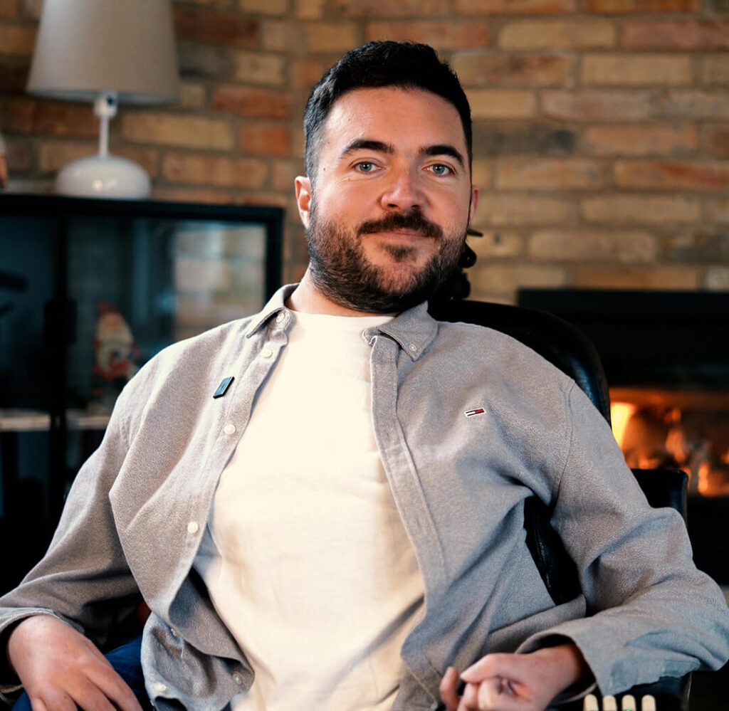 Portrait of a man with short dark hair and beard, sitting in a power wheelchair in front of a brick wall and a lit fireplace. He smiles gently and is wearing a light gray shirt over a white T-shirt.