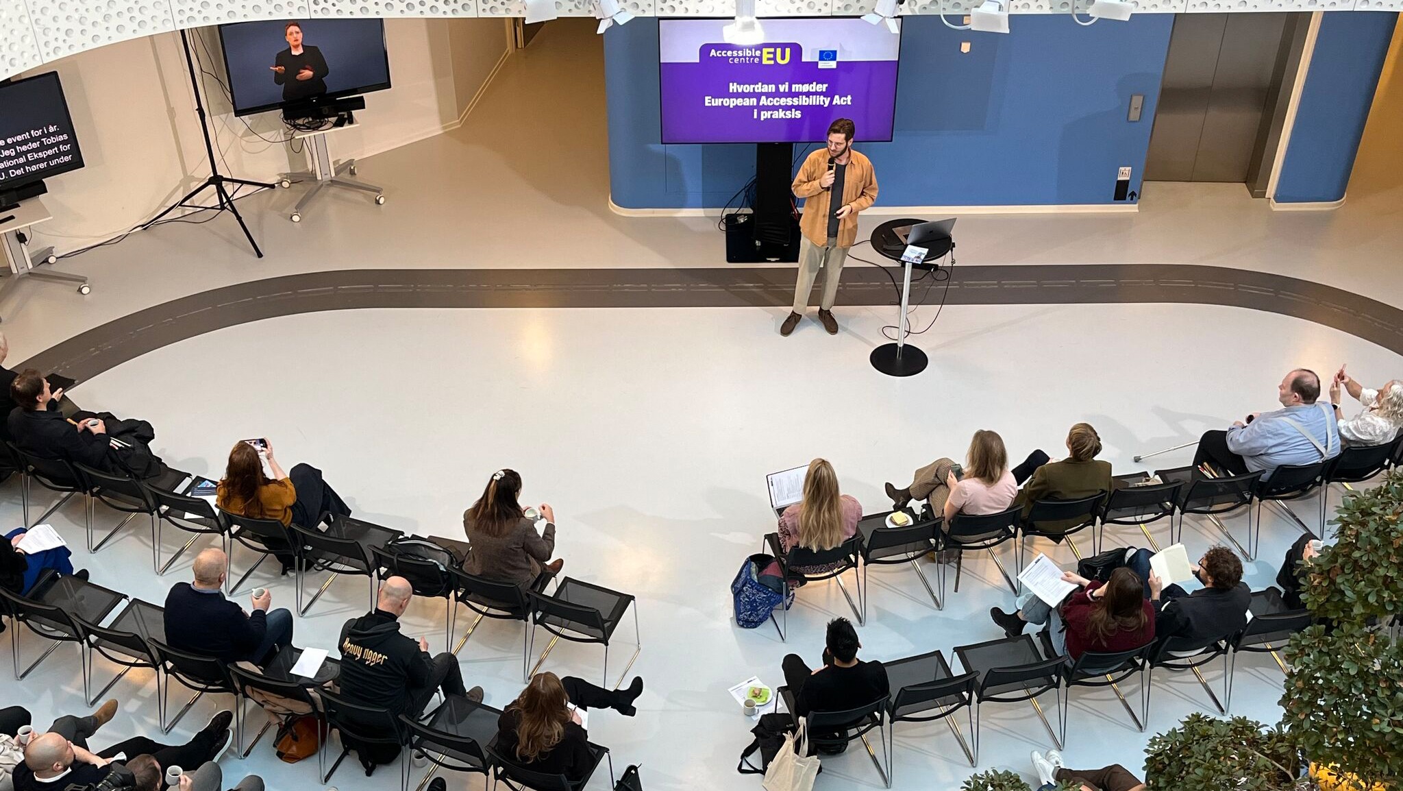 Seen from the balcony, Tobias speaks on a stage in front of an audience at the Danish Disability Organisations’ House. He stands at a small podium with a microphone, and behind him a large screen displays the title “How we meet the European Accessibility Act in practice”. The audience is seated in rows of chairs in a bright, open atrium hall. To the left, a screen shows a sign language interpreter and captions. Camera and audio equipment is set up around the room.