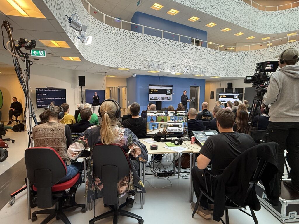 Publikum sidder i et stort, lyst atrium og følger et oplæg, mens tegnsprogstolkning og skrivetolkning vises på skærme. Bagest arbejder teknikere med kameraer, lyd og skærme, der viser både taler og tolke. Oplægsholderen er Lars Holm Sørensen, der demonstrerer skærmlæser-software for publikum.