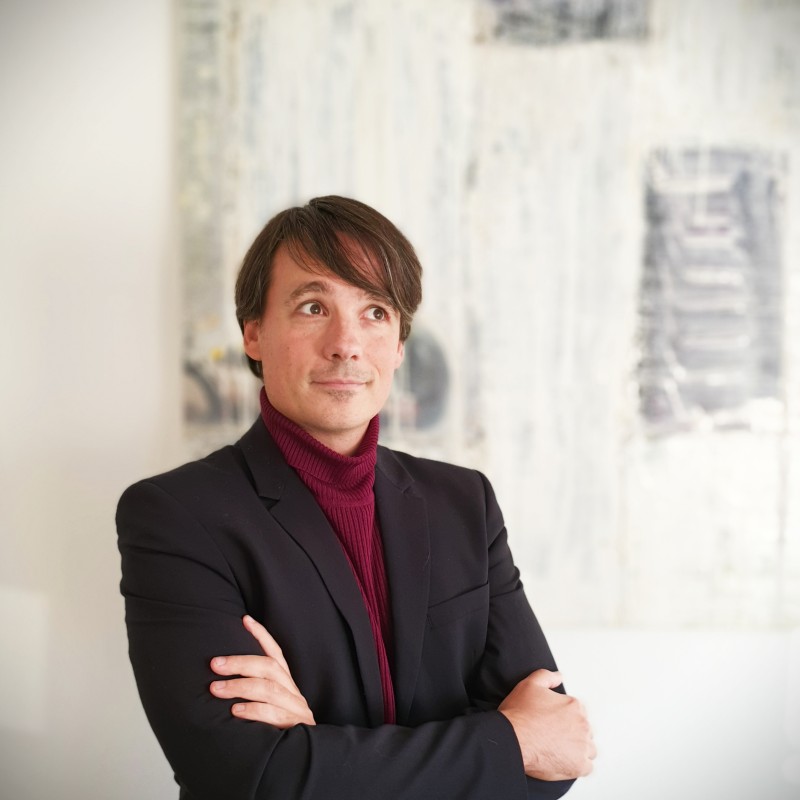 Portrait photo of a man with dark hair wearing a black blazer and a burgundy turtleneck, standing with arms crossed in front of a light wall with abstract art in the background.