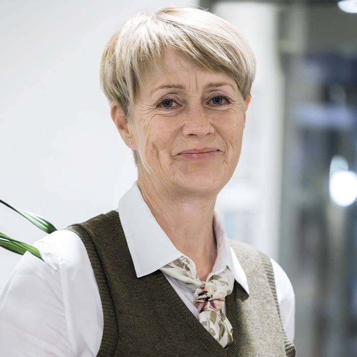 Portrait photo of a woman with short, light hair and a slight smile, looking directly into the camera. She is wearing a white shirt, a patterned scarf around her neck, and a brown vest. The background is a bright indoor setting with a blurred office area and a green plant in the background.