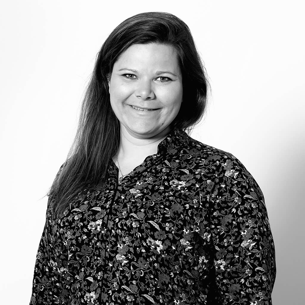 Black-and-white portrait photo of a woman with long dark hair smiling at the camera. She is standing slightly turned at the shoulders but looking straight ahead. She is wearing a dark shirt with a dense floral pattern.