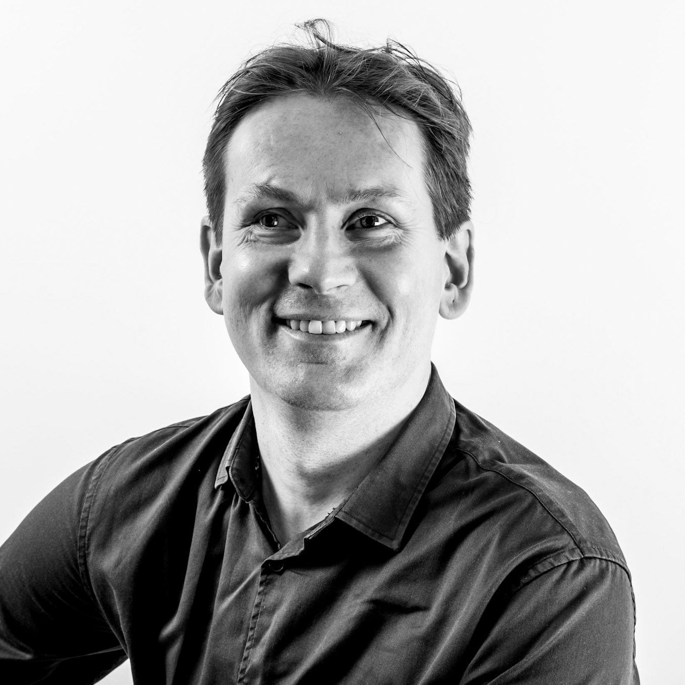 Black-and-white portrait of a smiling man with short hair looking directly into the camera. He is wearing a dark shirt, and the background is light and simple.