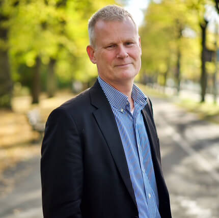 Portrait photo of a man with short blond hair wearing a dark blazer and a blue striped shirt, standing on a sunlit avenue with trees and a blurred background.