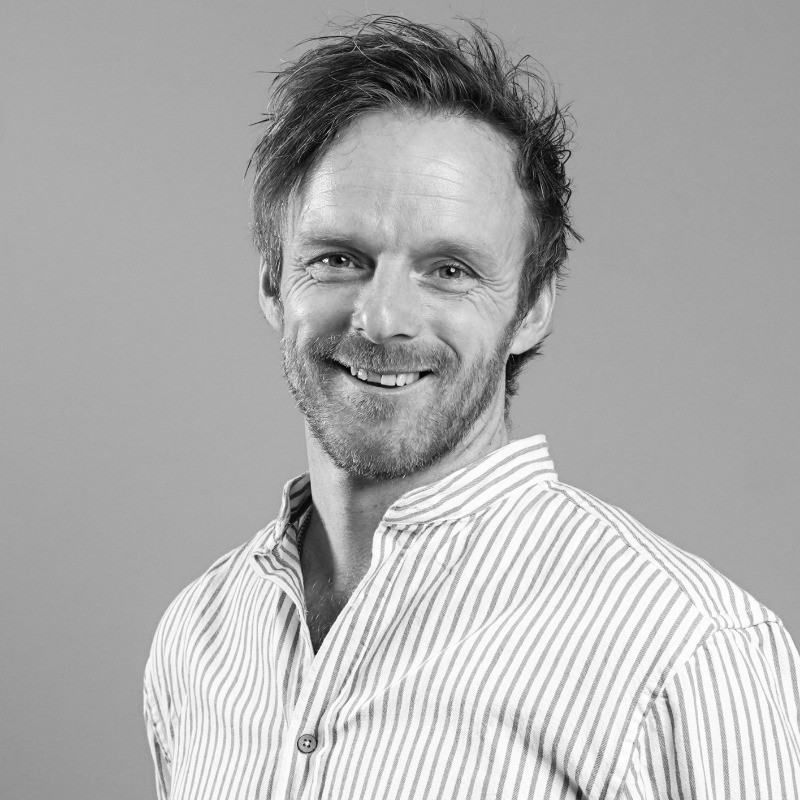 Black-and-white portrait of a smiling man with short, slightly messy hair and stubble. He is turned slightly to the side and looks into the camera. He is wearing a light shirt with narrow vertical stripes, and the background is light and simple.