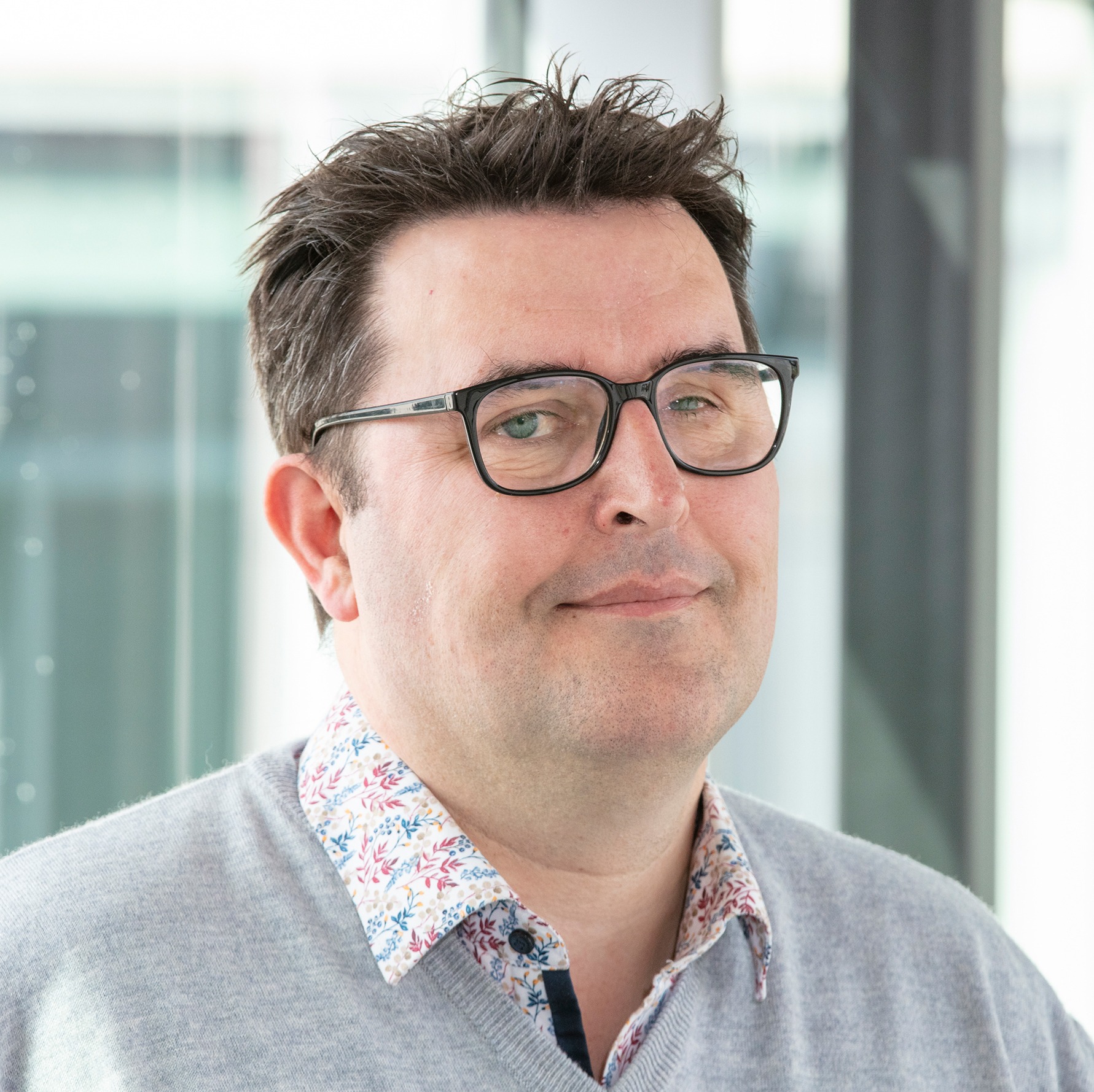 Portrait of a man with short dark hair and glasses, wearing a light grey sweater over a shirt with a colourful pattern. He looks calmly and with a slight smile at the camera. The background is bright and blurred.