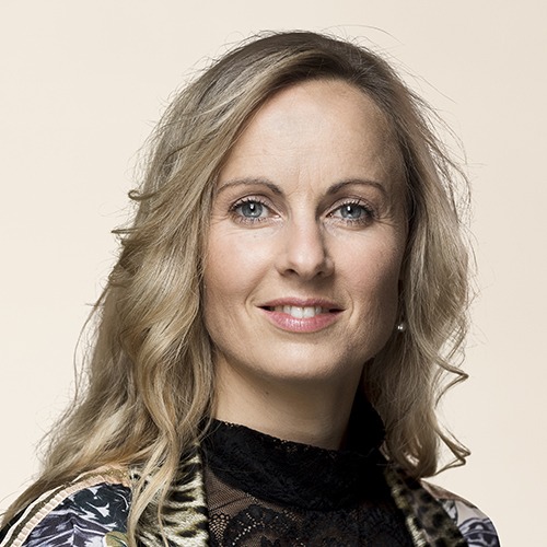 Portrait photo of a woman with light, shoulder-length hair and blue eyes. She looks into the camera and smiles slightly. She is wearing a dark blouse with a lace collar and a patterned jacket.