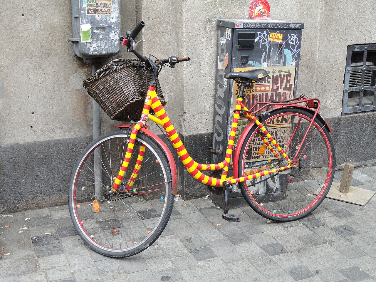 En rød og gulstribet cykel med kurv står op ad en graffitidækket elboks i København.